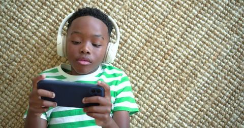 African american boy lying on jute rug listening with headphones and playing on smartphone