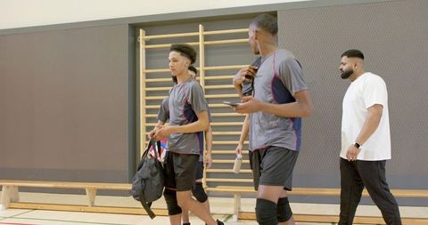 Diverse male team walking through indoor gym with coach, duffel bags and phones