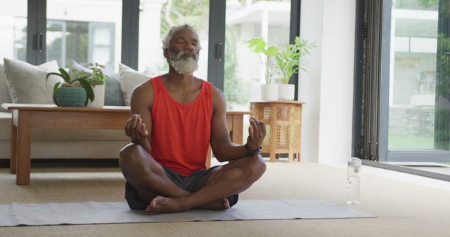 Senior man sitting cross-legged on yoga mat in bright living room, meditating peacefully. Suggest for articles on home workout routines, relaxation techniques, meditation practices for seniors, health and wellness concepts, or promoting mindfulness and well-being.