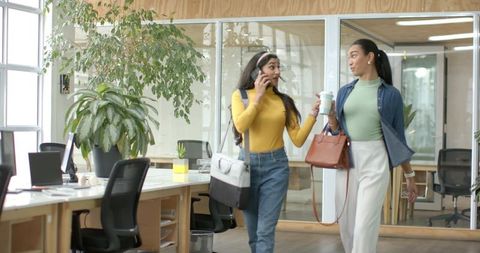Female colleagues walking through coworking office holding coffee while talking on phone