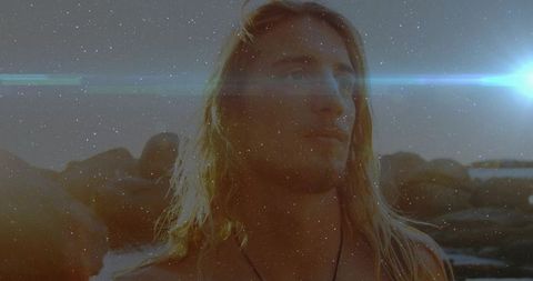Shirtless blond man standing on rocky shoreline gazing toward horizon during golden hour
