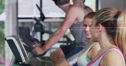 Smiling women chatting while using cardio machines in bright gym with tech reflections