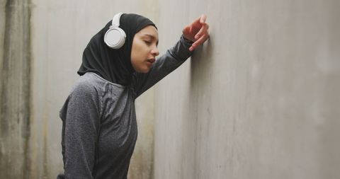 Tired athlete in hijab with headphones resting against wall