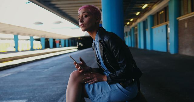 Young woman with pink hair sitting at train station holding smartphone. Wearing denim dress and black leather jacket. Vibe is modern and trendy, perfect for ads related to fashion, travel, urban living, communication, or young adult lifestyles. Useful for social media, blogs, websites, and promotional materials targeting young, urban demographics.