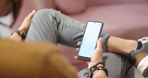 Casual man sitting on pink sofa holding smartphone with legs crossed and modern bracelets