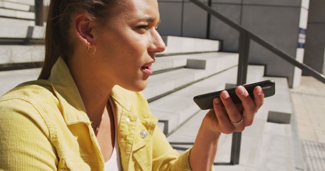 Woman using voice assistant feature on smartphone while sitting outdoors on steps. Ideal for illustrating casual technology use, communication, and modern lifestyles. Can be used for marketing technologies, mobile apps, or lifestyle articles.