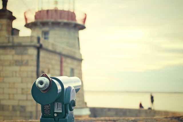 This image shows a green telescope mounted near a lighthouse overlooking the sea. The cloudy sky and soft lighting suggest evening or early morning. Useful for travel blogs, marine navigation articles, tourism advertisements, or inspirational outdoor prints.