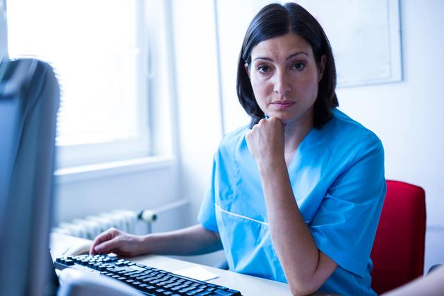 Female doctor sitting at desk in medical office, wearing blue medical uniform. She is looking at the camera with a serious expression, hand resting on chin. Computer and office equipment visible. Ideal for use in healthcare, medical practice, hospital, and professional work environment contexts.