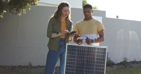 Young professionals installing solar panel and checking wiring with smartphone outdoors