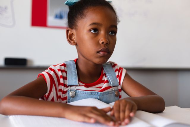 Young African American girl attentively studying braille in a classroom, highlighting inclusive education and the importance of learning tools for students with disabilities. Ideal for educational materials, special needs awareness campaigns, and inclusive learning resources.