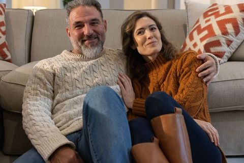 Mature couple cuddling on floor beside sofa wearing knit sweaters and boots, cozy home