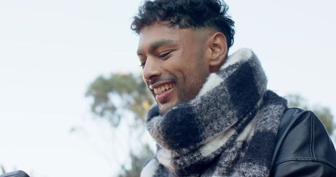 Young Asian man smiling while checking smartphone, wearing scarf and leather jacket