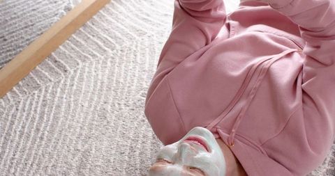 Young woman relaxing with facial mask lying on textured rug wearing pink hoodie