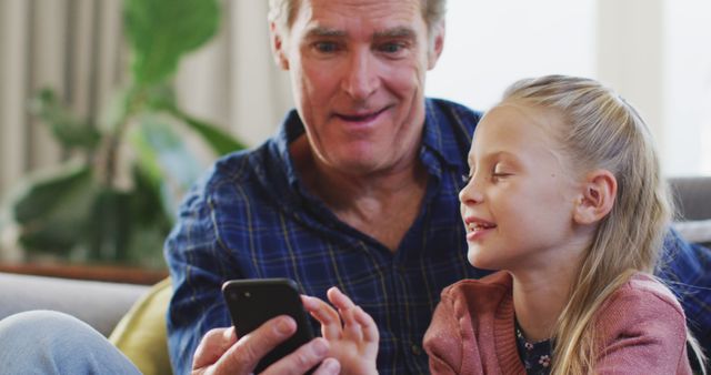 A cheerful grandfather is sitting with his smiling granddaughter at home, both looking at a smartphone. Ideal for advertisements about family time, technology use in everyday life, and intergenerational bonding. Suitable for use in promotions for mobile devices, family-oriented services, and home-related products.