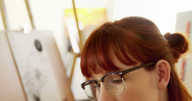 A young Caucasian woman with red hair and glasses is focused on her work in an art studio, with copy space. Her creative expression suggests she might be an artist deeply engaged in crafting her artwork.