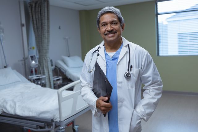 Mature male doctor standing confidently with a clipboard in a hospital ward. Ideal for use in healthcare, medical, and hospital-related content. Can be used in articles, brochures, and websites focusing on medical professionals, patient care, and hospital environments.