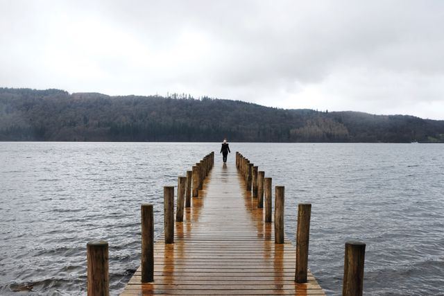 Solitary Figure Walking on Wooden Pier Over Placid Lake on Cloudy Day - Download Free Stock Images Pikwizard.com
