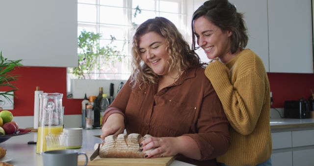 Two women bonding while preparing breakfast in a modern kitchen. One woman is cutting bread while the other looks on, smiling and supporting. Perfect for use in lifestyle blogs, cooking websites, or advertisements promoting kitchen products or breakfast foods.