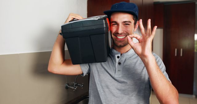 Young male plumber confidently carrying a toolbox while smiling and giving the OK sign in a modern kitchen. Ideal for advertisements and publications featuring home services, reliable handyman professionals, and positive customer-worker interactions.