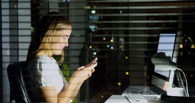 Woman sitting at desk engaging in tasks on her smartphone. Scene illuminated by computer screen and office lights. Useful for themes on late work nights, remote work, corporate life, technology in business, or communication in professional environments.