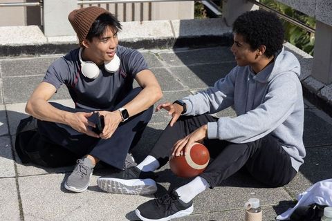 Diverse teens chatting on urban rooftop holding football, smartphone and headphones