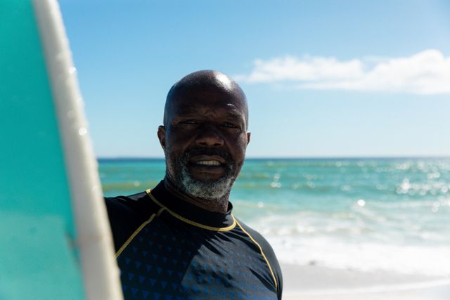 This image depicts a senior African American man standing on a beach with a surfboard, highlighting an active and adventurous lifestyle. Ideal for use in content related to retirement, fitness, outdoor activities, and holiday promotions. Perfect for promoting healthy living and active aging.