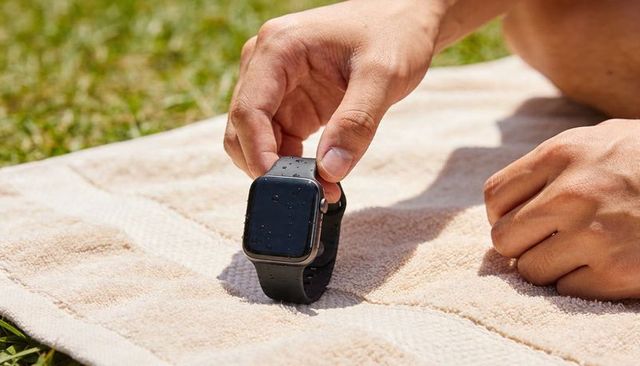 Hand adjusting wet smartwatch on towel outdoors with water droplets and sunlit grass