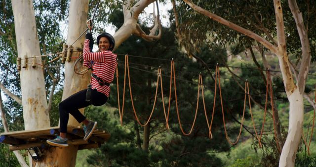 Woman smiling while participating in rope course adventure surrounded by trees in a scenic forest. Ideal for content related to outdoor activities, adventures, forest getaways, summer camps, team-building exercises, and promoting active and healthy lifestyles.