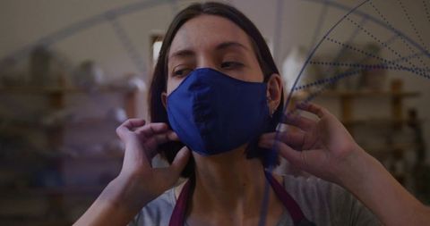 Artisan woman adjusting blue cloth face mask in wood workshop, closeup with maroon apron