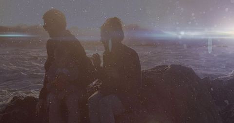 Silhouetted family sitting on rocky shore holding bundled baby at sunset with ocean flare