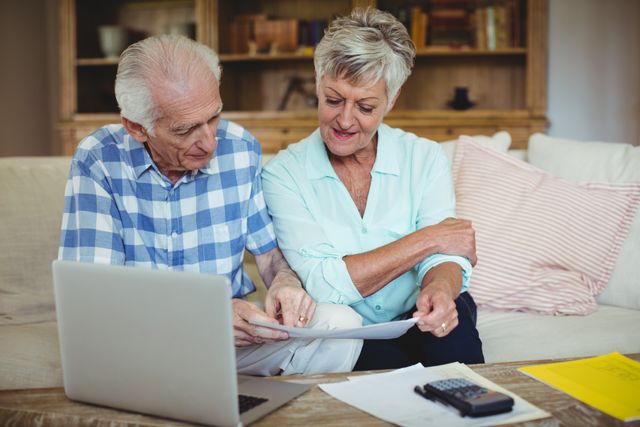 Senior couple sitting on a couch in a living room checking bills together with a laptop. Great for content on retirement planning, family budgeting, financial advice for seniors, or promoting financial products for elder demographics.