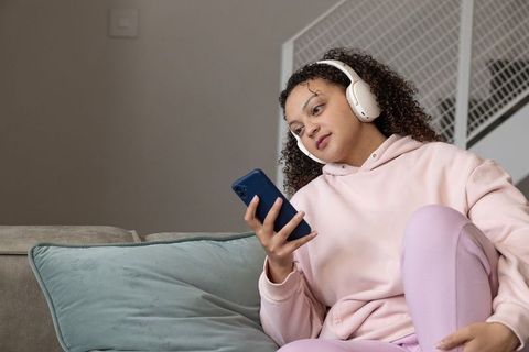 African american woman relaxing on sofa with headphones and smartphone in pastels