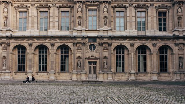 Historic building with classical architecture, showing intricate stone facades and detailed statues. Two people are sitting on a bench in the cobblestone courtyard. Valuable for use in travel blogs, architectural articles, historical sites, educational materials, and brochures.