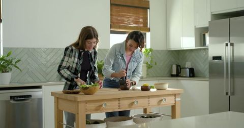 Diverse female friends preparing meal at modern kitchen island grinding herbs together