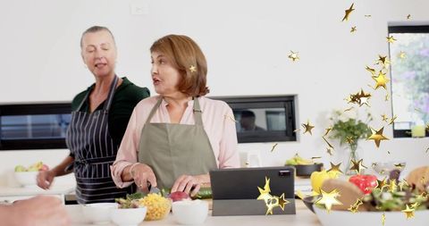 Two women chopping vegetables and preparing fresh salad at kitchen island with tablet
