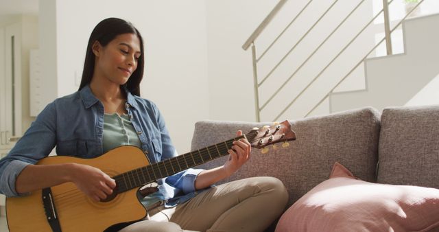 Young woman sitting on a sofa in a modern home interior, playing an acoustic guitar and enjoying a relaxed moment. Great for concepts of relaxation, enjoying music at home, leisure activities, and peaceful lifestyle. Useful for websites and blogs focused on music, hobbies, home living, and modern interiors.