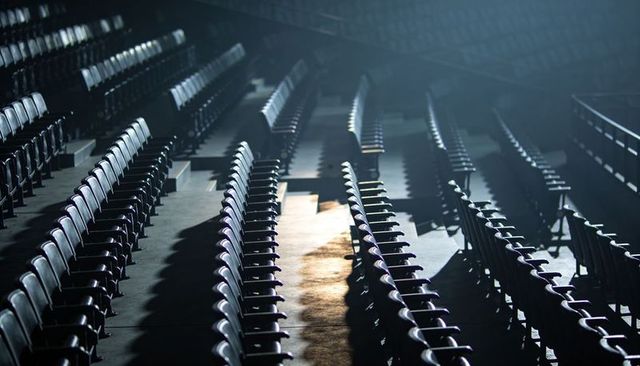 Empty tiered stadium seats casting long shadows along aisle under moody lighting