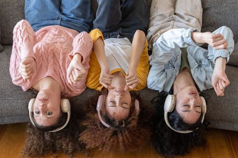 Three young women lying upside down on couch wearing wireless headphones, relaxing and listening to