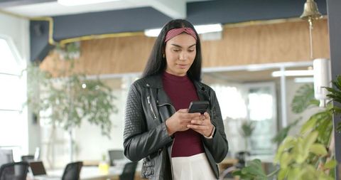 Asian woman texting on smartphone in coworking space, leather jacket and pink headband