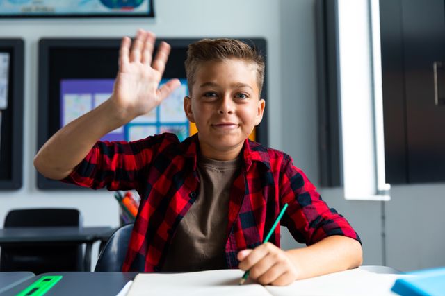 Portrait of happy caucasian boy sitting at desk, smiling and raising hand in elementary school class. Education, childhood and learning concept.