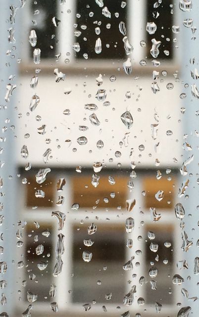 Raindrops on the window create a calming and reflective scene, with the blurred building in the background adding depth and context. This can be used for themes of rainy weather, seasons, mood, or ambiance in both personal and commercial projects such as blogs, websites, magazines, or even in residential or office space decor.
