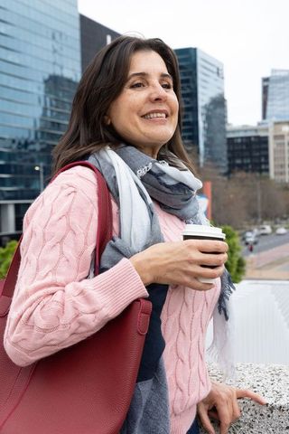 Smiling commuter enjoying coffee on city balustrade wearing pink sweater and red tote