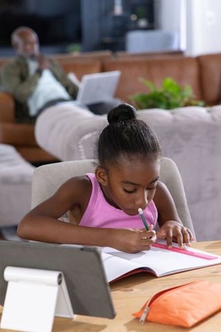 African american girl studying homework at table while father works remotely on couch