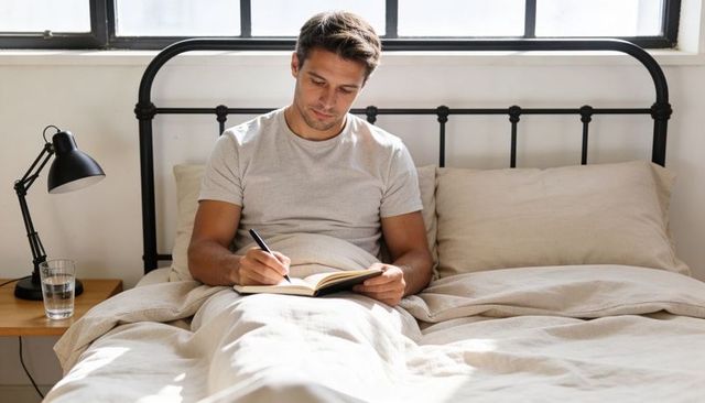 Man Writing in Bed Morning Journaling with Notebook and Pen in Minimalist Sunlit Bedroom