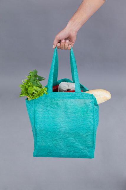 Close-up of a hand holding a reusable grocery bag filled with fresh produce including leafy greens and a baguette. Ideal for use in articles or advertisements promoting eco-friendly shopping, sustainability, healthy lifestyles, and organic food choices.