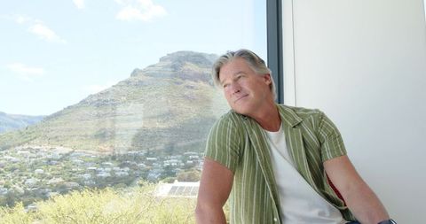Mature man gazing through window at mountain valley while sitting on modern home ledge