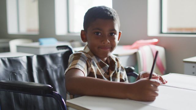 A young male student sitting in a classroom, appearing engaged and content while writing at a desk. This depiction emphasizes classroom inclusion and education for diverse and differently-abled students. Useful for materials promoting inclusive classroom environments, educational resources, and school diversity awareness campaigns.