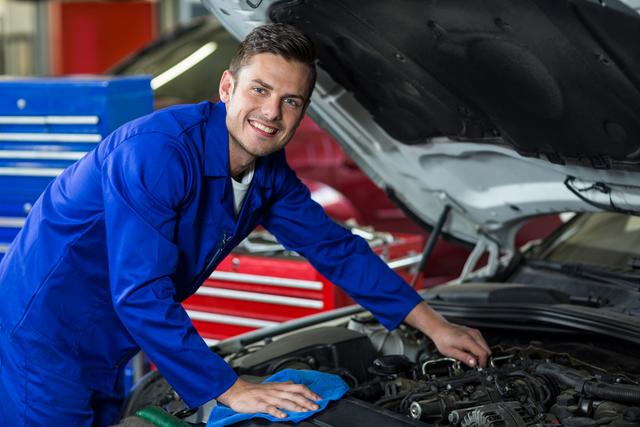 Mechanic in blue uniform servicing car engine in repair garage. Ideal for illustrating automotive repair services, professional mechanics, and vehicle maintenance. Can be used in advertisements for car repair shops, automotive service centers, and technical training programs.