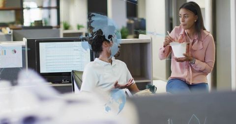 Two coworkers collaborating over takeout in open-plan office with analytics on monitors