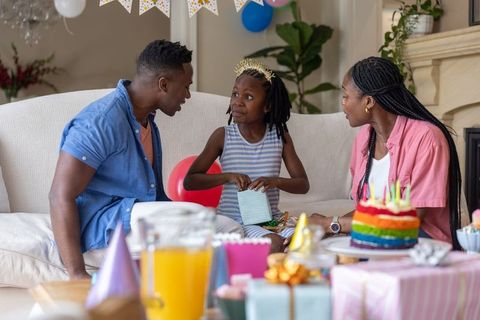 African american family celebrating daughter birthday at home opening gifts by rainbow cake
